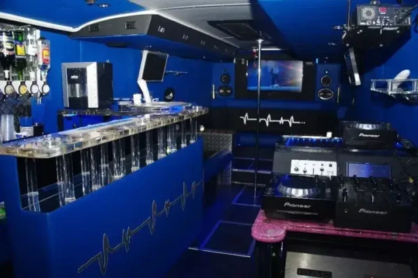 An image of the interior of a Prague XXXL Party Bus with vibrant blue lighting. It features a bar area with liquor dispensers, a TV screen, seating benches, and a DJ setup with Pioneer equipment. Perfect for a stag do, the walls and bar counter have heartbeat line designs, contributing to the festive atmosphere.