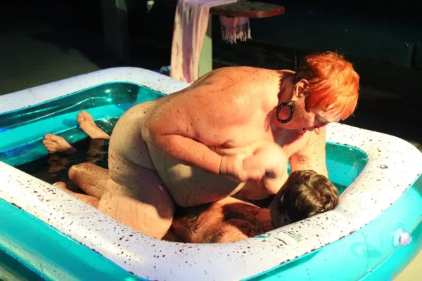 Two adults are inside an inflatable pool. One person with short red hair and large earrings is on top of another person with short hair. The scene appears intimate and intense, both partially submerged in water, reminiscent of wild Prague stag fun. The background appears dark.