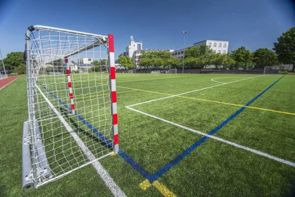 A soccer field with bright green artificial turf, yellow and white boundary lines, and a goalpost with a white net and red markings at one end. In the background, there are buildings and trees under a clear blue sky—perfect for some pre-game action before your Prague Football Match kicks off.