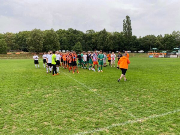 A group of soccer players from two teams in different uniforms gather on a grassy field, with trees and spectators in the background. A person in an orange outfit is walking towards the group, perhaps reminiscing about their recent Prague Football Match while the sky appears cloudy.