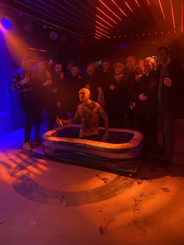 A group of people standing in a dimly lit club during a wild stag party. One person is in a small inflatable pool in the center, covered in dark liquid and holding a hammer, while the others stand around holding drinks. The red and blue lighting adds to the dramatic Prague stag fun atmosphere.