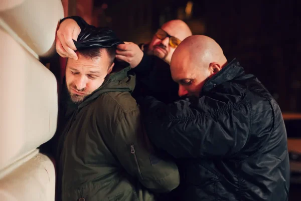 Three men in dark clothing restrain a man in a green jacket at night against a building wall. The restrained man, part of a Prank Stag Arrest & Strip, appears distressed, while the two other men seem focused, with one securing the man's hands behind his back and the other pressing him against the wall.