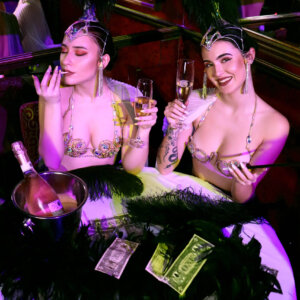 Two women dressed in ornate costumes with feathered headpieces sit at a table. One woman is smoking while the other holds a champagne flute, smiling. The table, reminiscent of a lively stag party in Prague, is adorned with dollar bills, black feathers, and an ice bucket with a bottle of Sexy VIP Darling Cabaret.